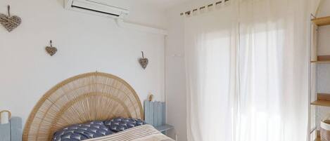 A bedroom with a bed, an air conditioner, a window with sheer curtains, and decorative heart-shaped wall hangings.