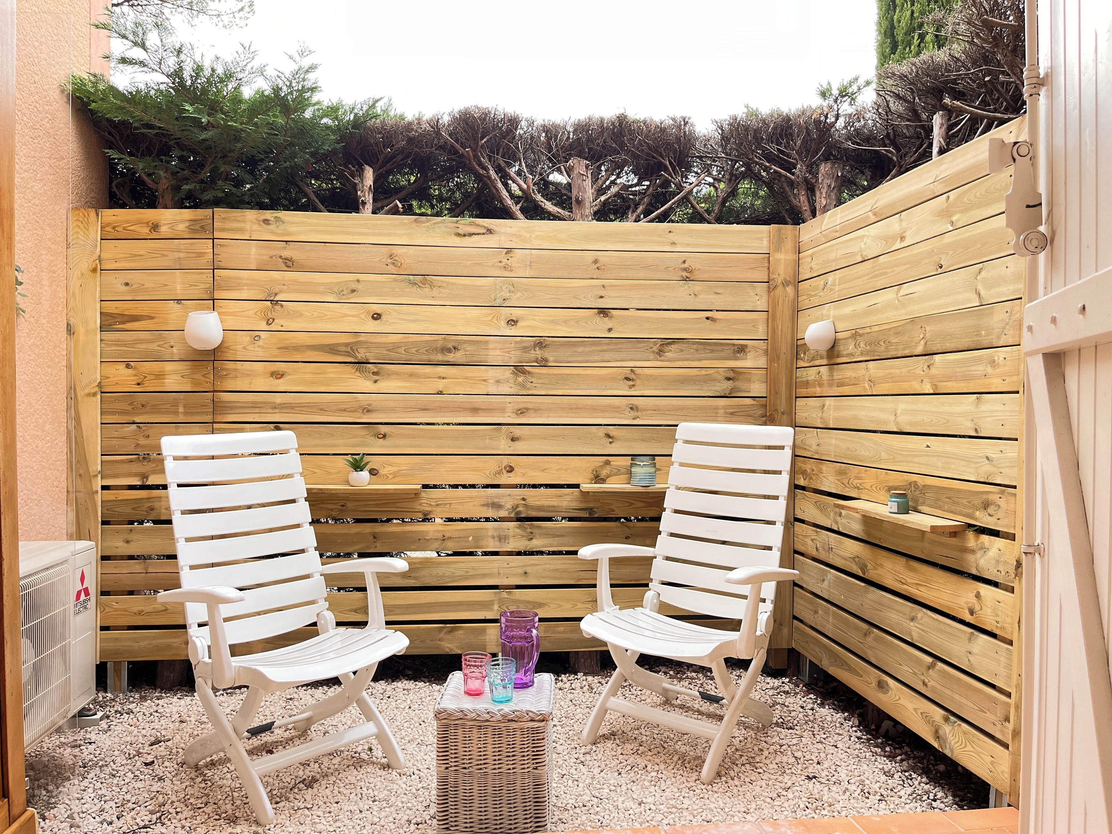 A wooden deck with two white chairs and a small table, surrounded by a wooden fence and greenery.