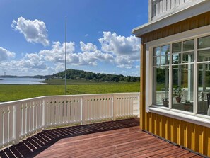 A wooden deck with a white railing, large windows, and a view of a green landscape and water. - 5 Star Holiday Home in Askeron (Hjaelteby)