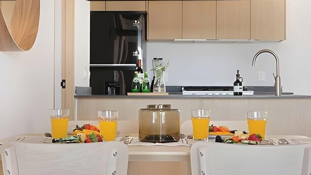 Une cuisine moderne avec des meubles de rangement en bois, un réfrigérateur noir et un évier. On y trouve deux chaises autour d’une table dressée pour deux personnes, avec un verre de jus d’orange et une petite salade devant chaque convive.