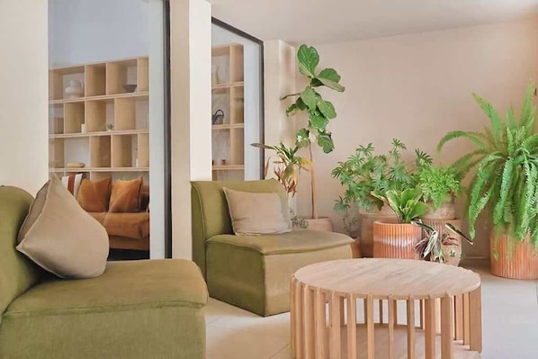 A living room with a sofa, armchair, and a round wooden coffee table. There are potted plants and a bookshelf in the background.