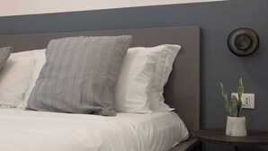A neatly made bed with white and gray striped pillows, a gray headboard, and a small round table with a plant beside the bed. - Cozy Trieste Historic Hideaway (Trieste)