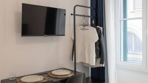 A hotel room with a flat-screen TV mounted on the wall, a clothing rack with a white shirt and a jacket, a dining table with a placemat and a glass, and a window with curtains. - Cozy Trieste Historic Hideaway (Trieste)