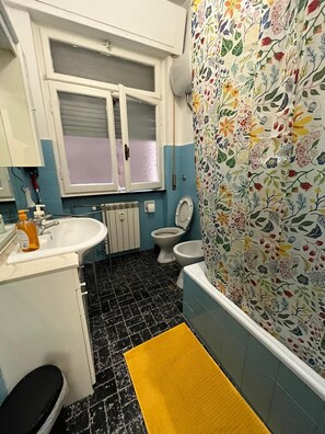 Een badkamer met een witte wastafel, twee toiletten, een badkuip met bloemenbehang en een geel badmatje.