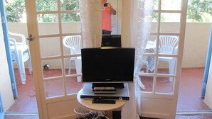 Una habitación con una televisión sobre una mesita, una silla blanca y una puerta que da a un espacio exterior con sillas blancas y suelo de baldosas.