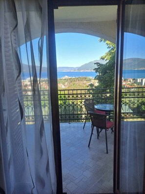 A balcony with a table and chairs, offering a view of a coastal landscape with buildings and greenery. - Apartment Sea View  3 (Tivat)