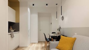 Eine moderne Küche mit weißen Schränken, ein Essbereich mit Tisch und Stühlen sowie ein Wohnzimmer mit einer Couch und einem gelben Kissen.