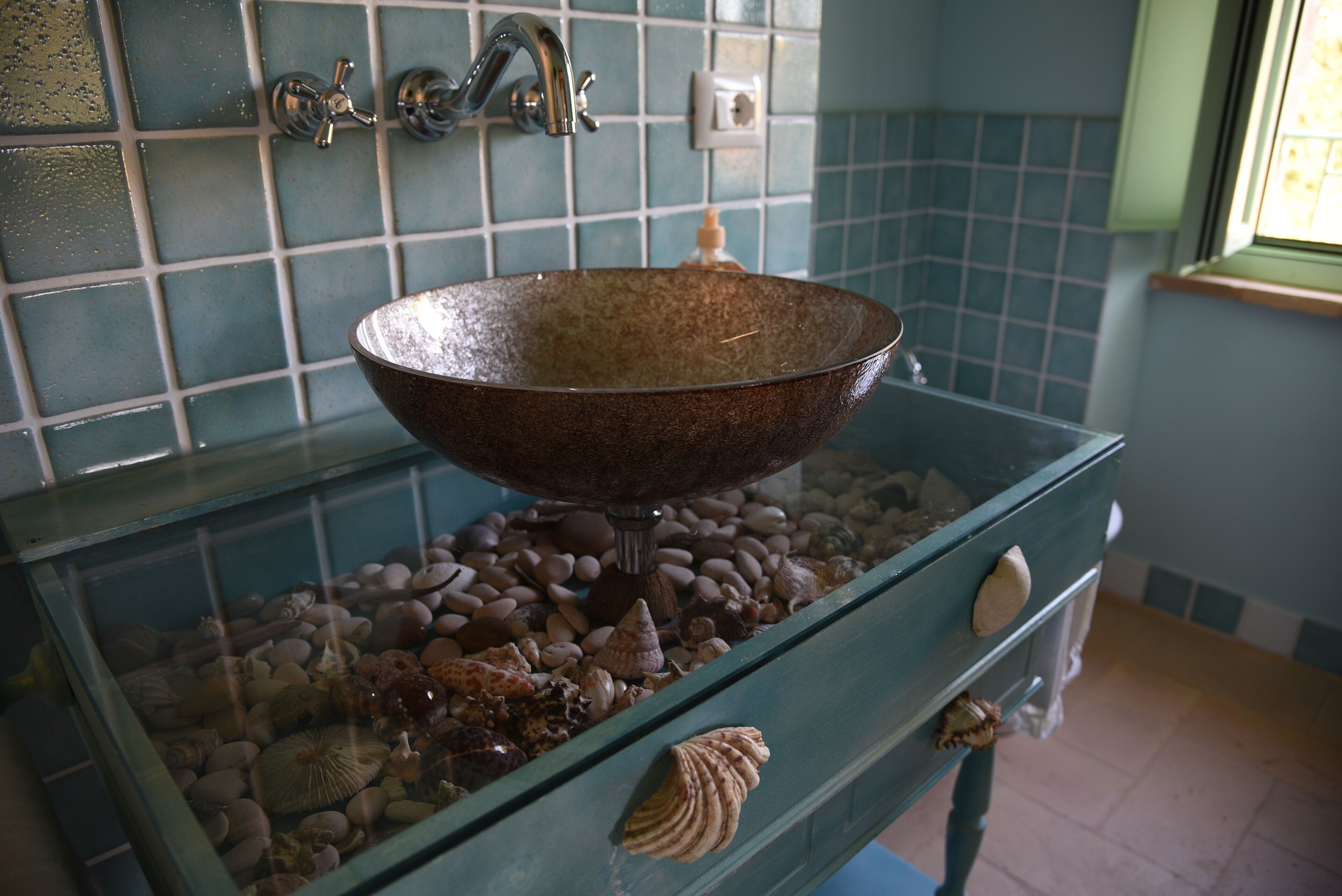 Un baño con lavamanos de vidrio, conchas decorativas y un salpicadero de azulejos.