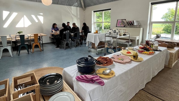 Un espacio interior amplio con una larga mesa dispuesta para una comida, rodeada de sillas y mesas.