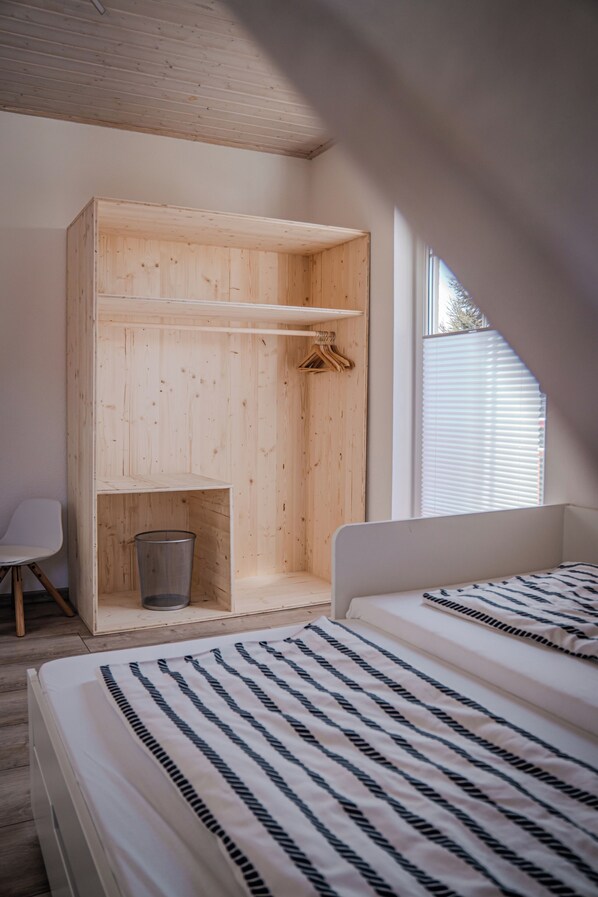 A bedroom with a wooden wardrobe, a small trash bin, a white chair, and a bed with striped bedding.