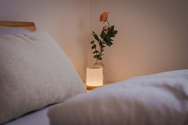 Una cama con ropa blanca, una mesita de noche con un florero que contiene una sola rosa, y una lámpara de noche encendida.