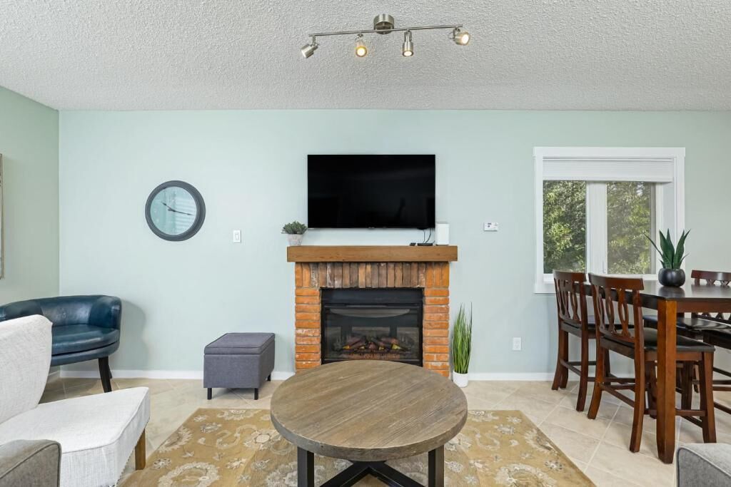 Un salon avec une cheminée, un téléviseur fixé au-dessus, une table à manger avec des chaises et une horloge au mur.