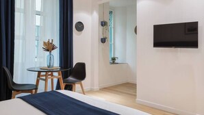 A modern hotel room with a bed, a TV mounted on the wall, a small table with chairs, and a view of the hallway. - Trieste Time Capsule (Trieste)