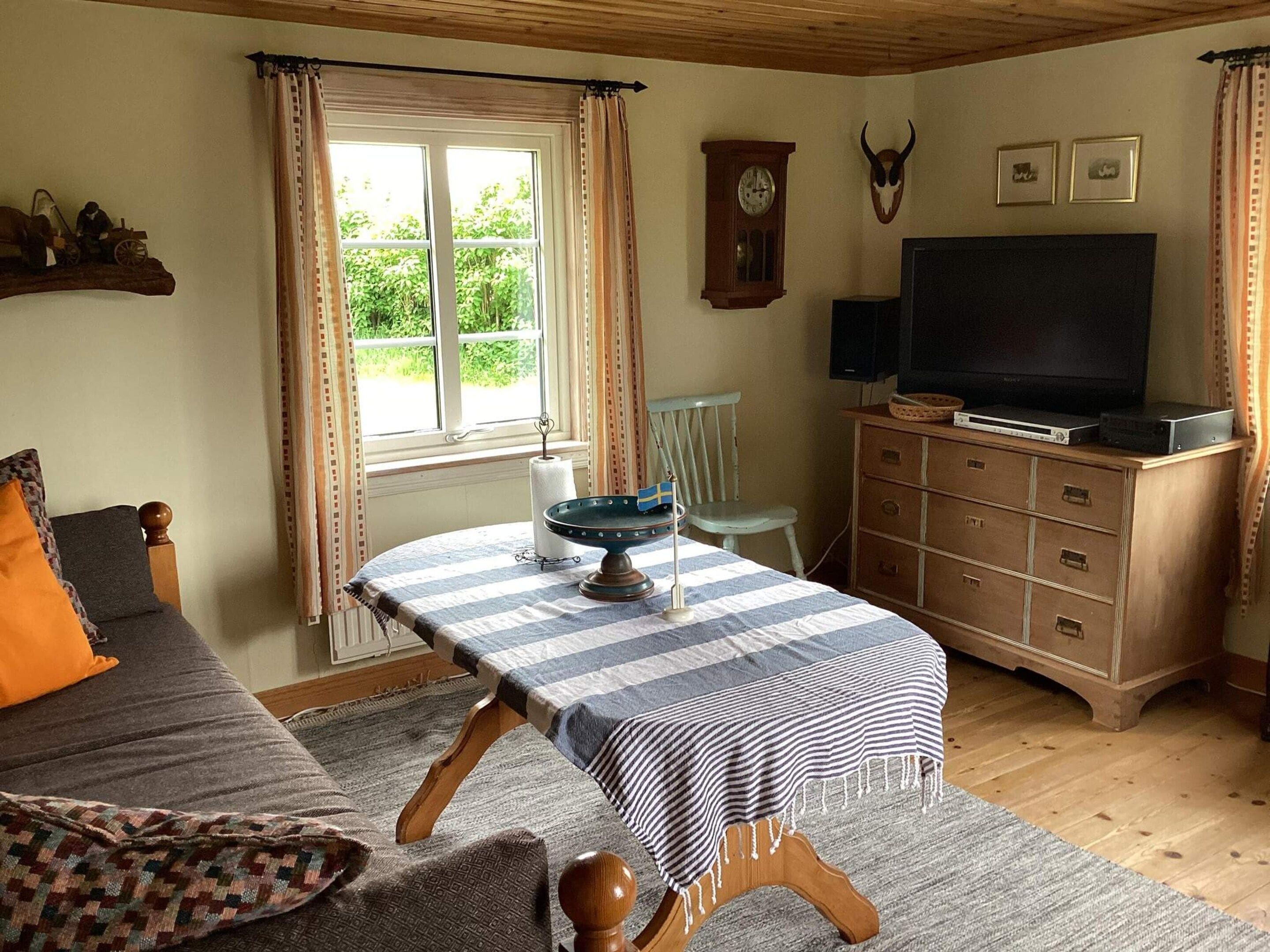 A cozy room with a bed, a table with a striped tablecloth, a television, a clock, and a wooden cabinet.