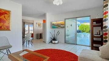 A modern living room with a red rug, a glass coffee table, a white sofa, and a TV mounted on the wall. There is a sliding glass door leading to a balcony with outdoor furniture.