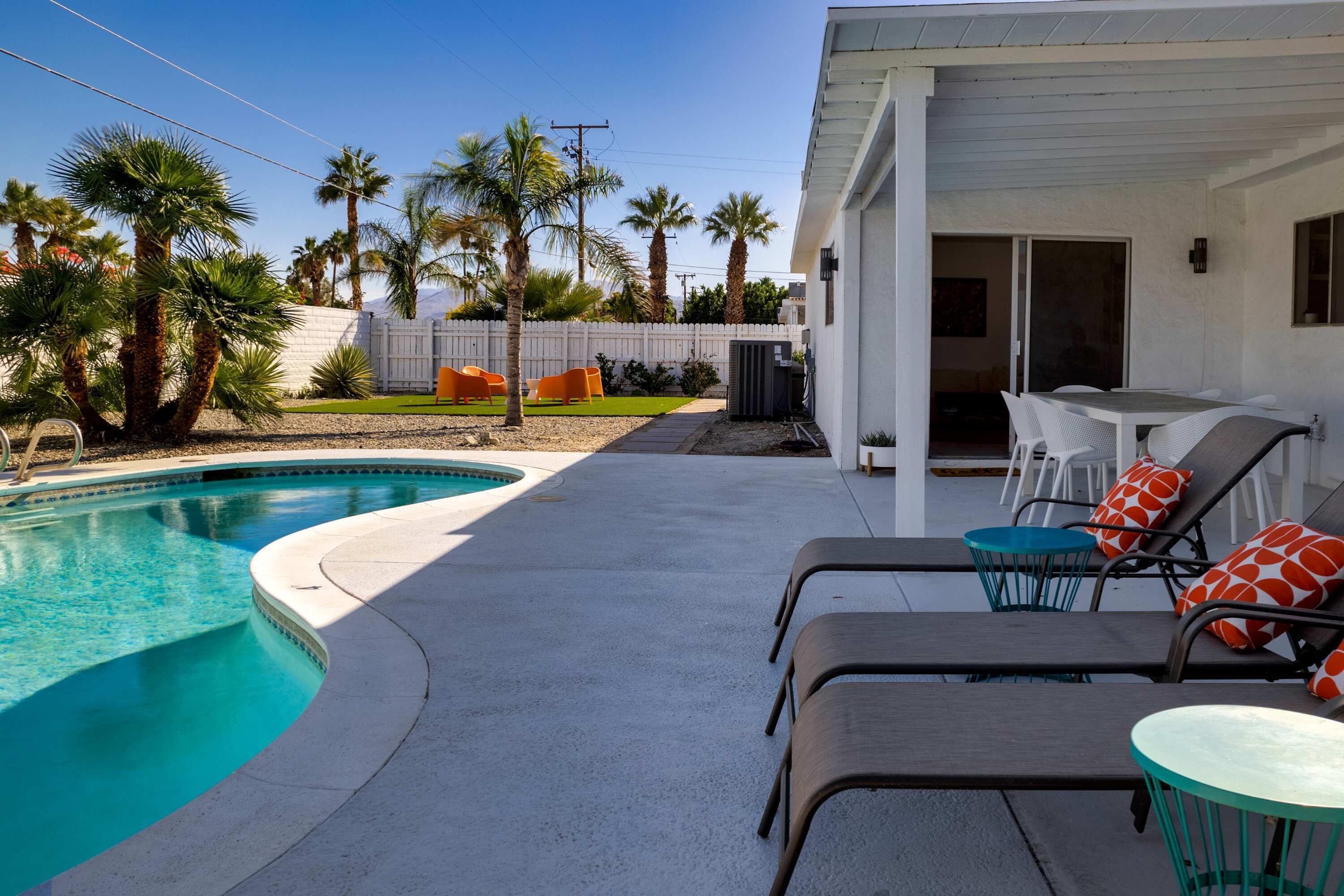 A pool area with lounge chairs and a table, surrounded by palm trees and a white fence.