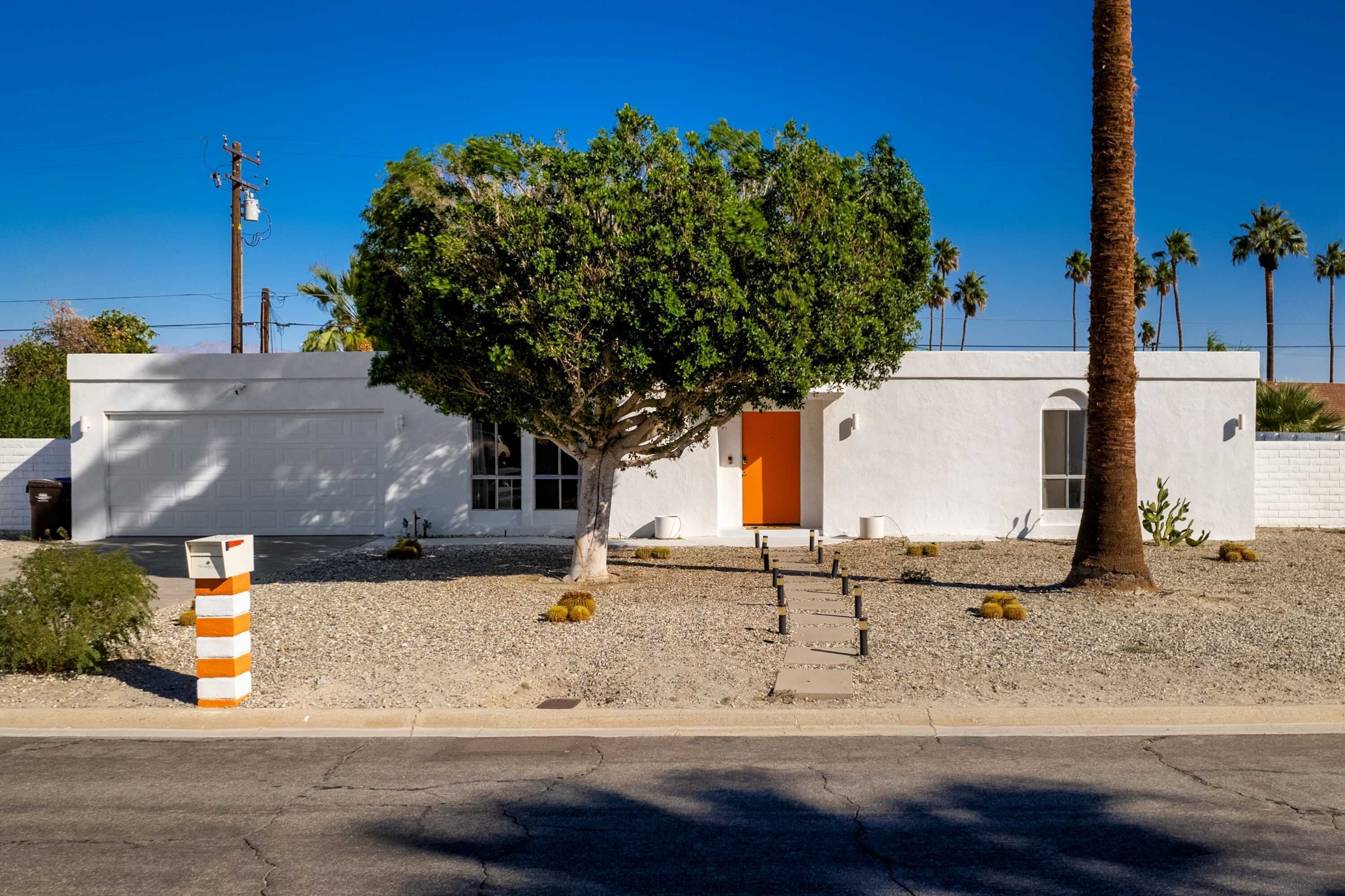 Un bâtiment blanc avec une seule porte orange, entouré de palmiers et surplombé d’un ciel bleu clair.