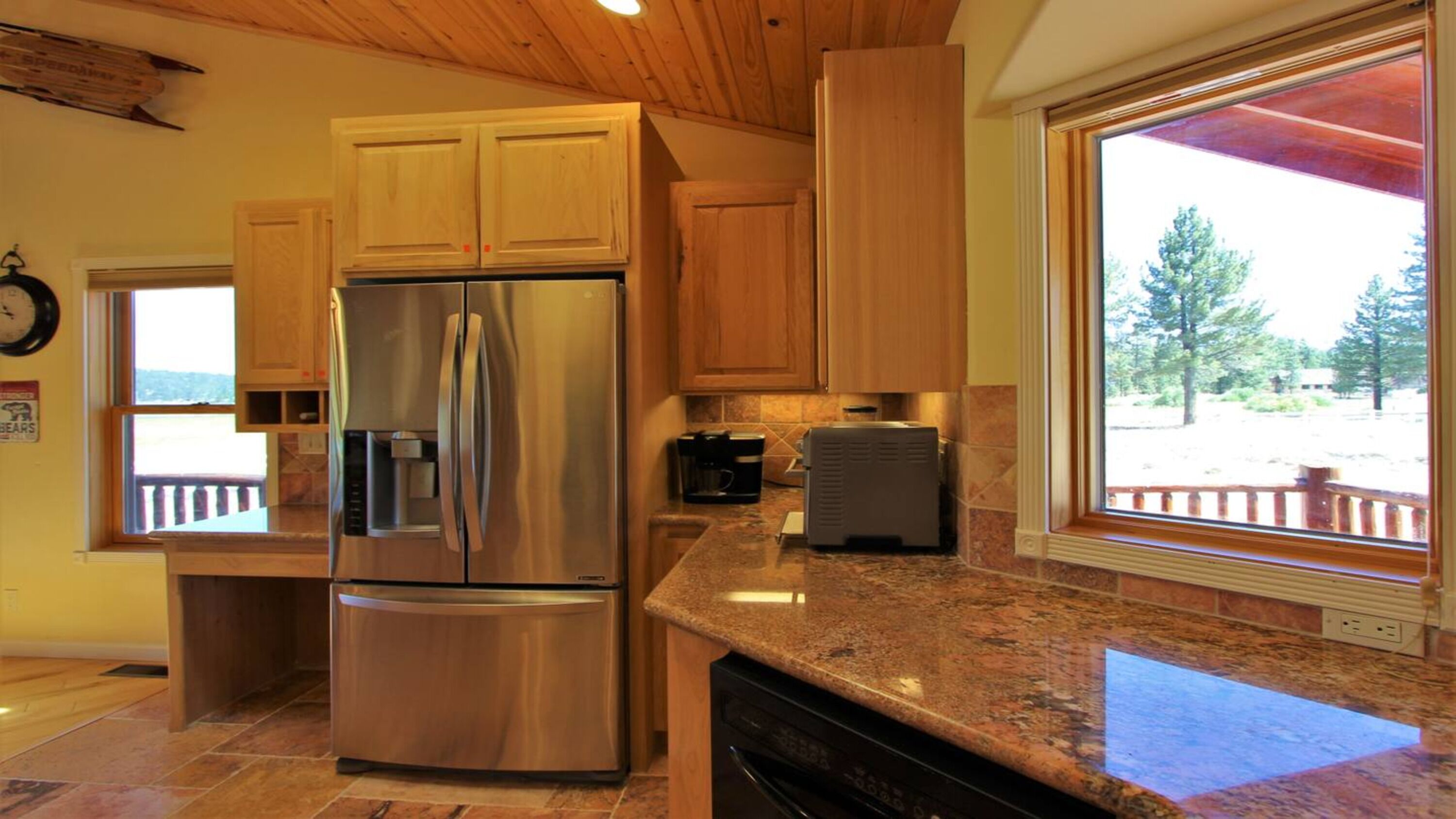 A kitchen with wooden cabinets, a stainless steel refrigerator, granite countertops, and a window with a view of trees.