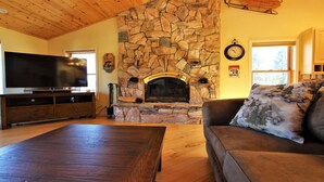 Un soggiorno con camino in pietra, televisore a schermo piatto, arredi in legno e un divano con cuscino decorativo.