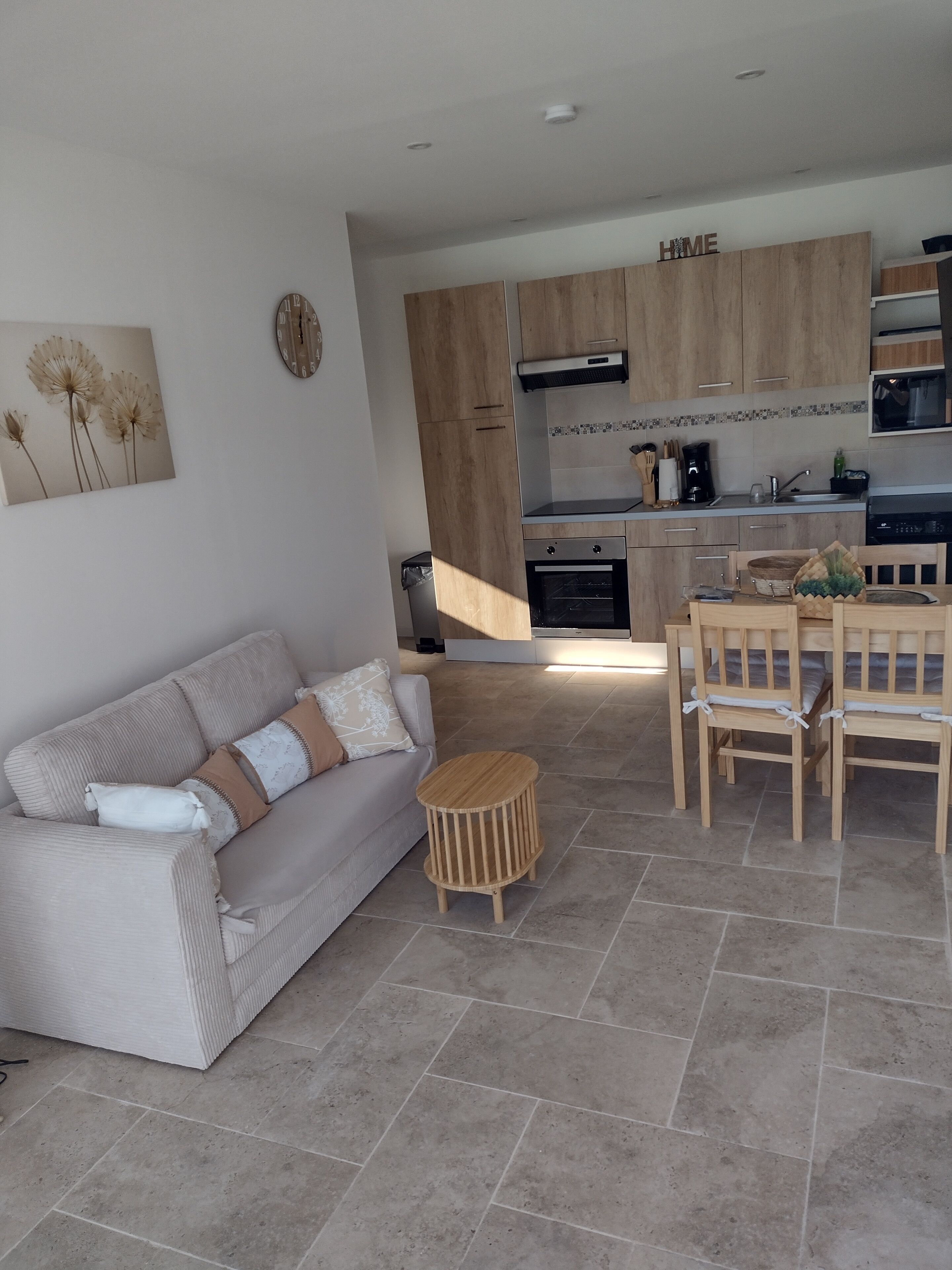 A modern kitchen with wooden cabinets, a dining area with a wooden table and chairs, and a living area with a sofa and a small wooden stool.