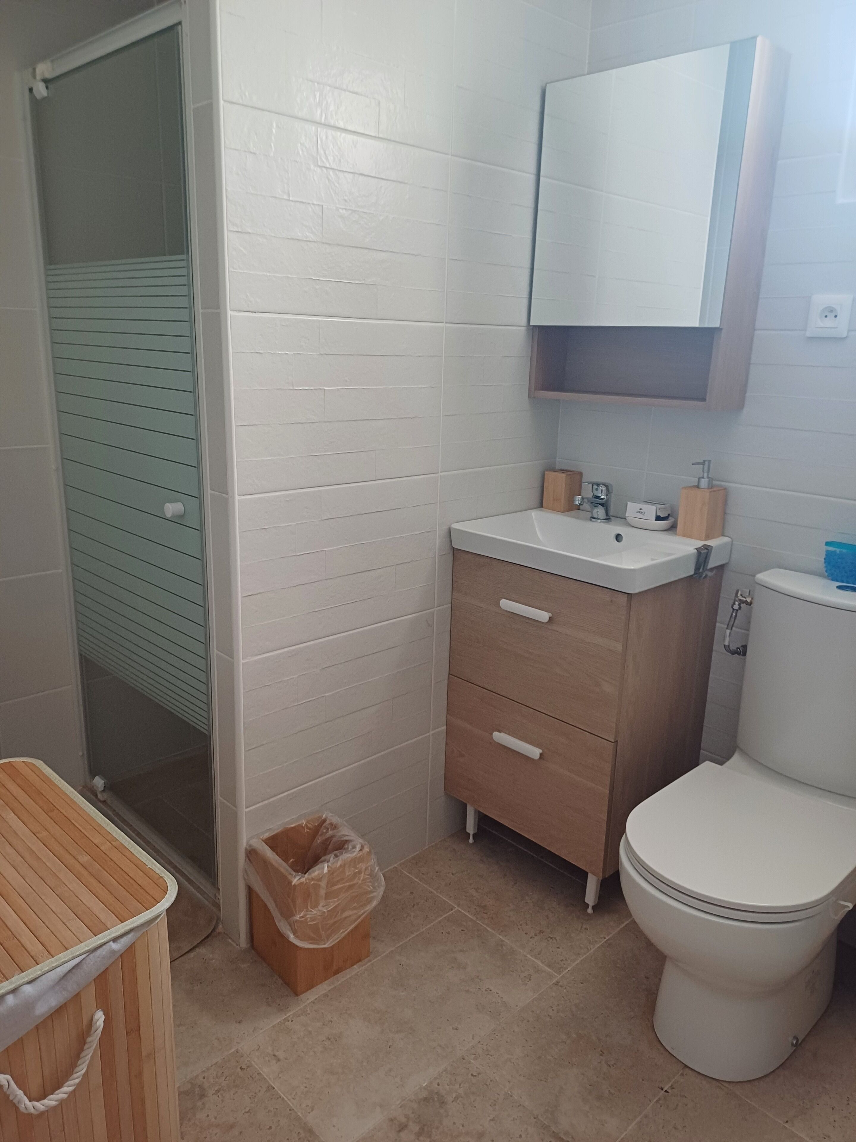 A bathroom with a white toilet, a sink with a wooden cabinet above, a trash bin, and a shower area with a glass door.