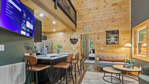 Un interior moderno con techo de madera, una zona de bar con taburetes, un sofá y un televisor que muestra servicios de streaming.