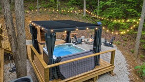A hot tub with a wooden deck and a pergola, surrounded by trees and string lights.