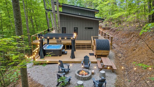 A cabin with a hot tub, fire pit, and deck seating surrounded by trees.