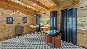 Interior de una cabaña de madera con un tapete de cuadros, un sillón colgante, una mesita con una lámpara y vistas al exterior con vegetación.