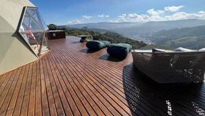 Una terraza de madera con cerramiento de vidrio, asientos verdes y vistas a colinas distantes.