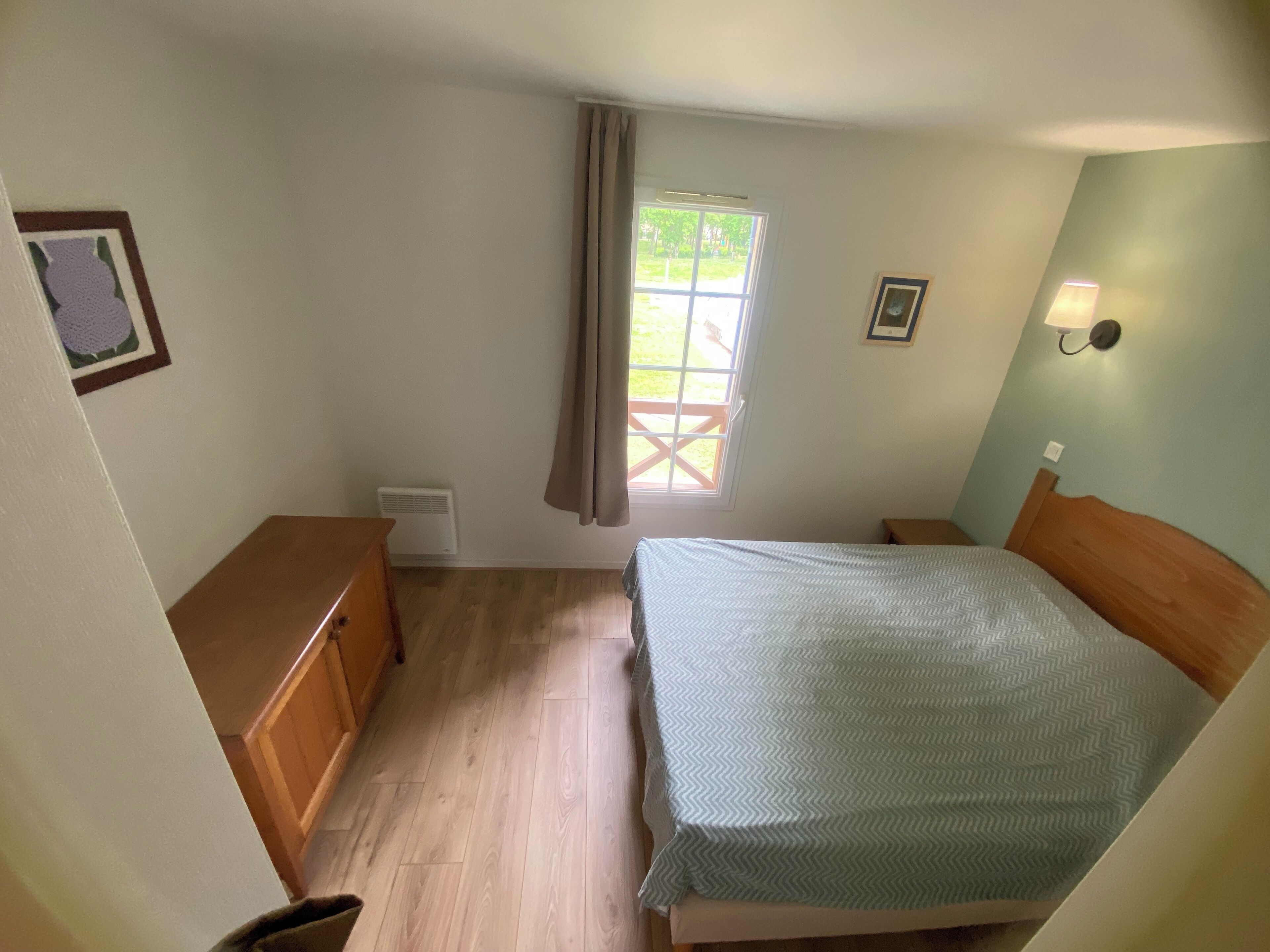 Ein Schlafzimmer mit Bett, Nachttisch, Kommode, einem Fenster mit Vorhängen und einer Wandleuchte.