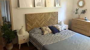 Ein Schlafzimmer mit einem Zierzargen an der Bettkopfteil, einem Bett mit einem geometrischen Muster auf der Bettdecke, Nachttischen, einer Kommode und einem an der Wand befestigten Regal mit gerahmten Bildern und einem Spiegel.