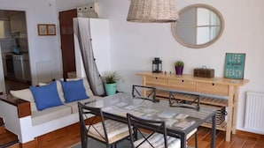Ein modernes Wohnzimmer mit einem Glastisch, einem Sofa mit blauen Kissen, einer Hängeleuchte aus Rattan, einem runden Spiegel und einer Holzkonsole mit dem Schild 'Life is good at the beach'.