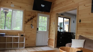 A wooden cabin interior with a television mounted on the wall, a door with a window, a white shelf, a wicker basket on a table, and a sofa with a patterned pillow.