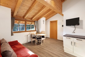A room with a wooden ceiling, a red sofa, a dining table with chairs, a TV mounted on the wall, and a door.