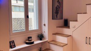 Un escalier avec des marches en bois et un mur blanc. On y voit deux fenêtres avec des stores, un climatiseur fixé au mur et un tableau encadré.