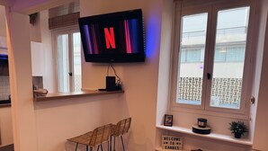Un salon moderne avec un téléviseur à écran plat affichant le logo de Netflix, un tabouret de bar en osier, une pancarte de bienvenue et une fenêtre donnant sur l’extérieur.