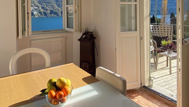 A dining area with a table, chairs, and a bowl of fruit. There are open windows with a view of mountains and a lake. - Laglio Cesare’s House Lake View (Laglio)
