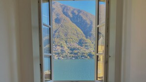 A room with a window overlooking a lake and mountains, a white chair, and a wooden floor. - Laglio Cesare’s House Lake View (Laglio)