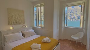A bedroom with a bed, two windows, a chair, and a painting on the wall. - Laglio Cesare’s House Lake View (Laglio)