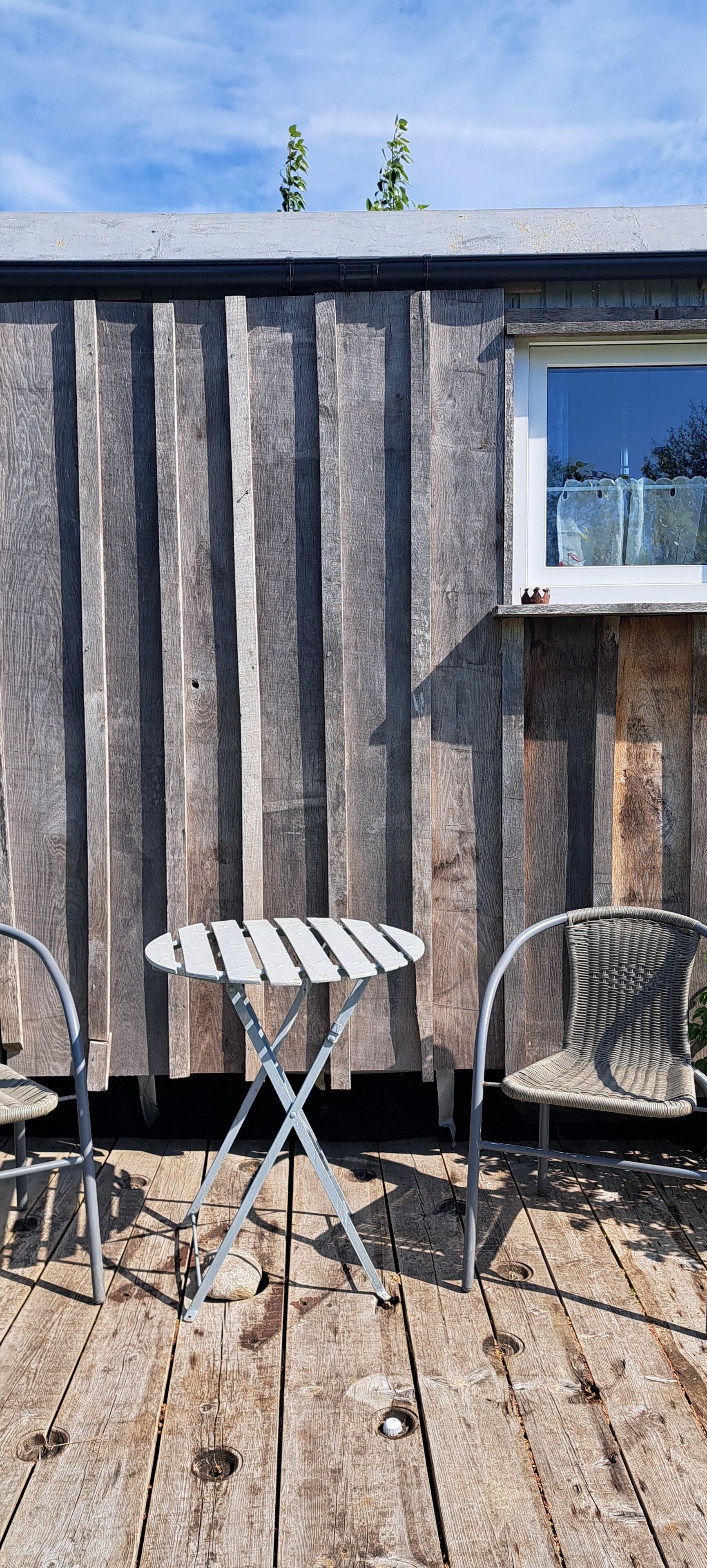 A wooden deck with a metal table and two chairs, a window with a view, and a wooden fence.