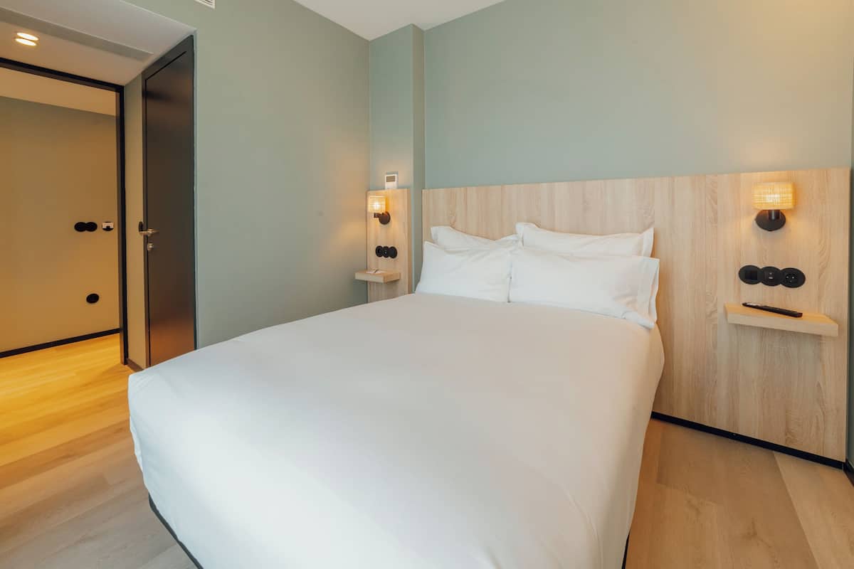 A modern hotel room with a large bed, wooden headboard, and wall-mounted lighting.