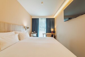 Habitación de hotel con una cama grande, un escritorio con silla, televisor montado en la pared y una ventana con cortinas.