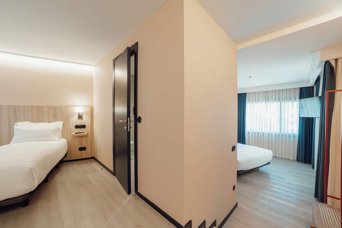 A hotel room with two beds, a bedside table with a lamp, a wardrobe, and a view of the outside through a window with curtains.