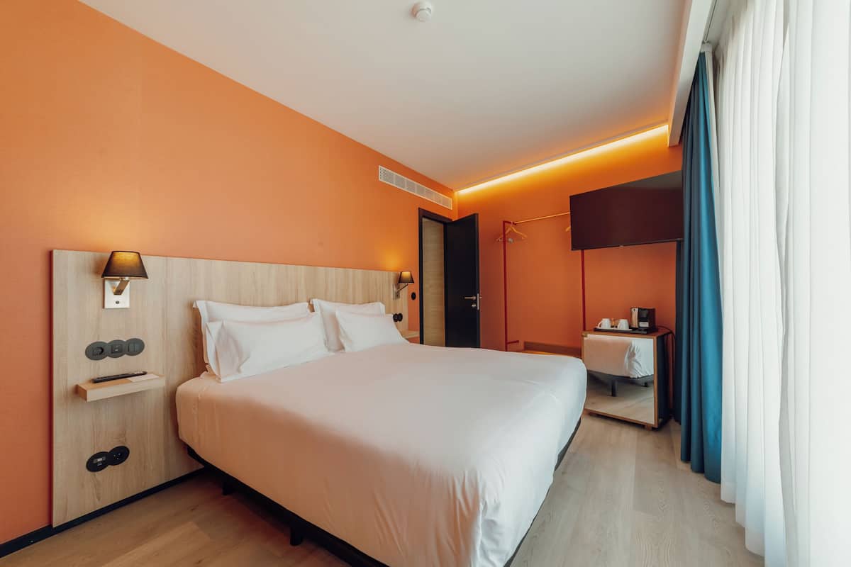 A hotel room with a large bed, a bedside table with a lamp, a wardrobe, a TV, and a bathroom with a shower and toilet.