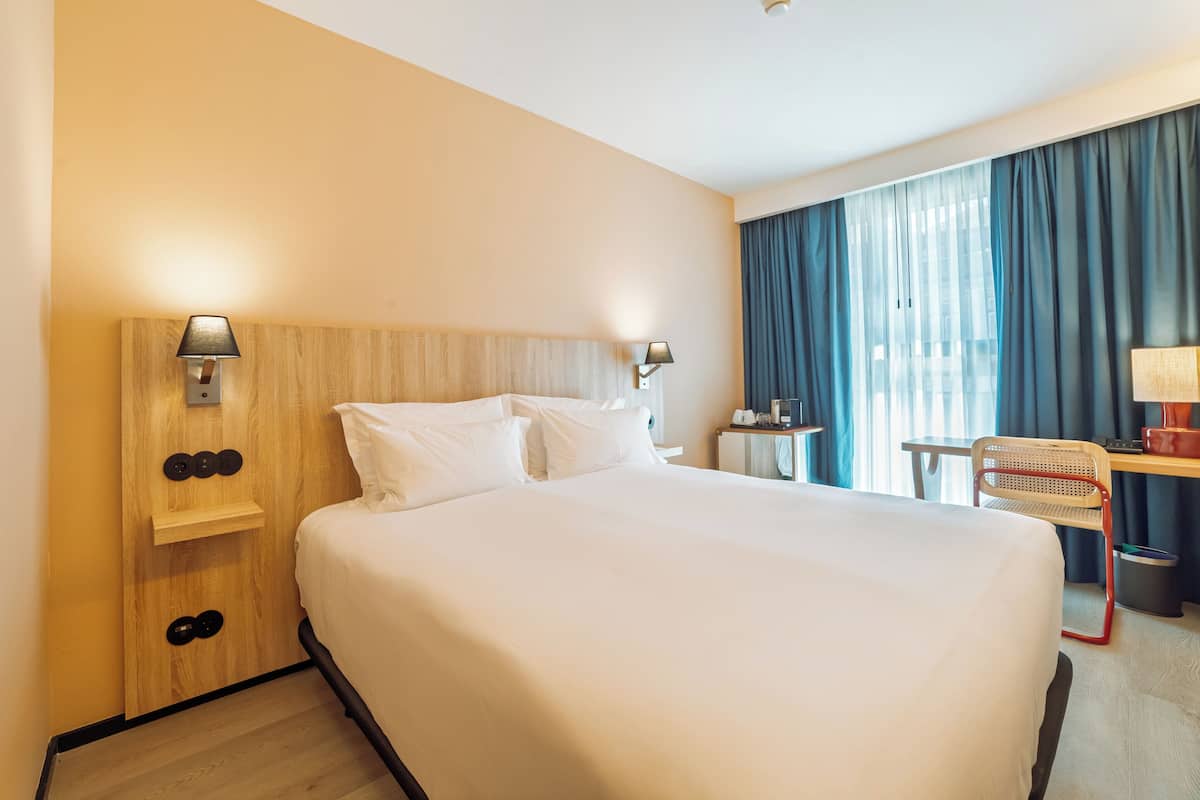 A hotel room with a large bed, wooden headboard, bedside lamps, a desk with a chair, and a view of the outdoors through a window with curtains.