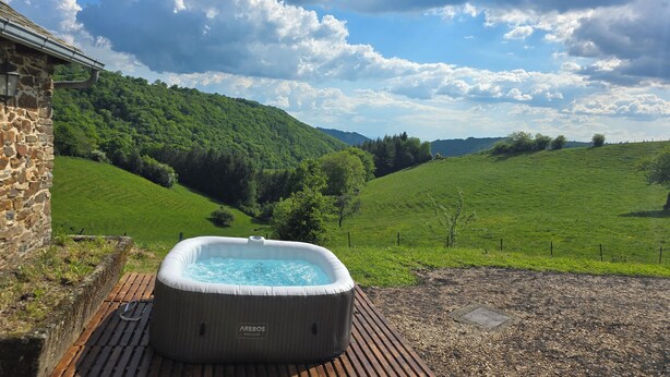 A hot tub on a wooden deck with a stone building and a lush green hillside in the background. - Authentic Aubrac farmhouse with Jacuzzi. A change of scenery guaranteed! (Montpeyroux)