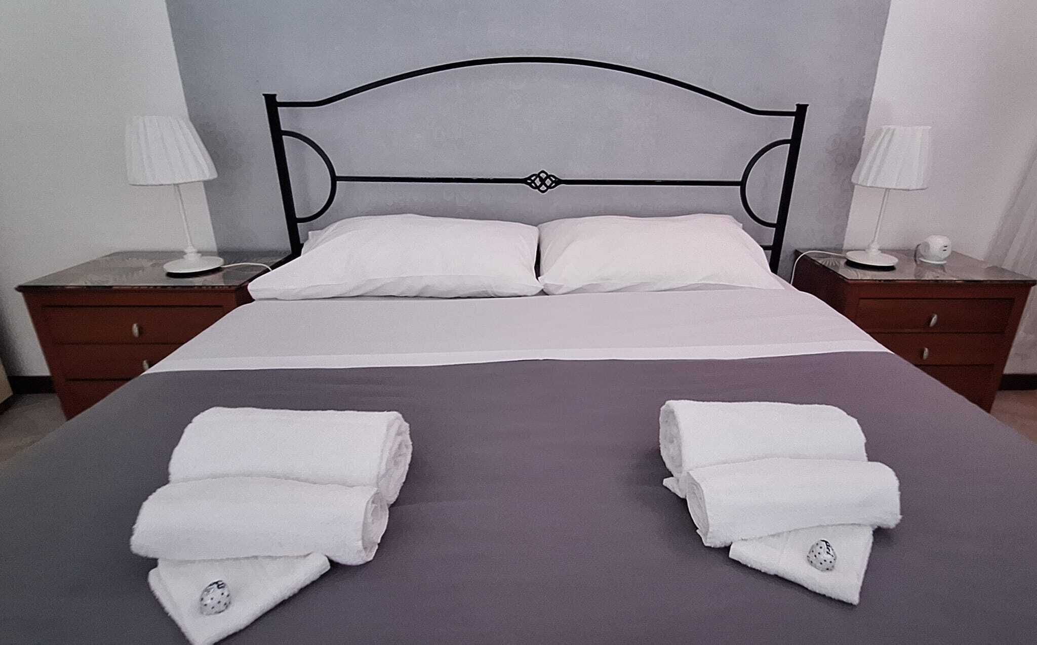 Un lit double avec une tête de lit en métal, du linge de lit blanc, deux tables de chevet avec des lampes et des serviettes pliées posées sur le lit.