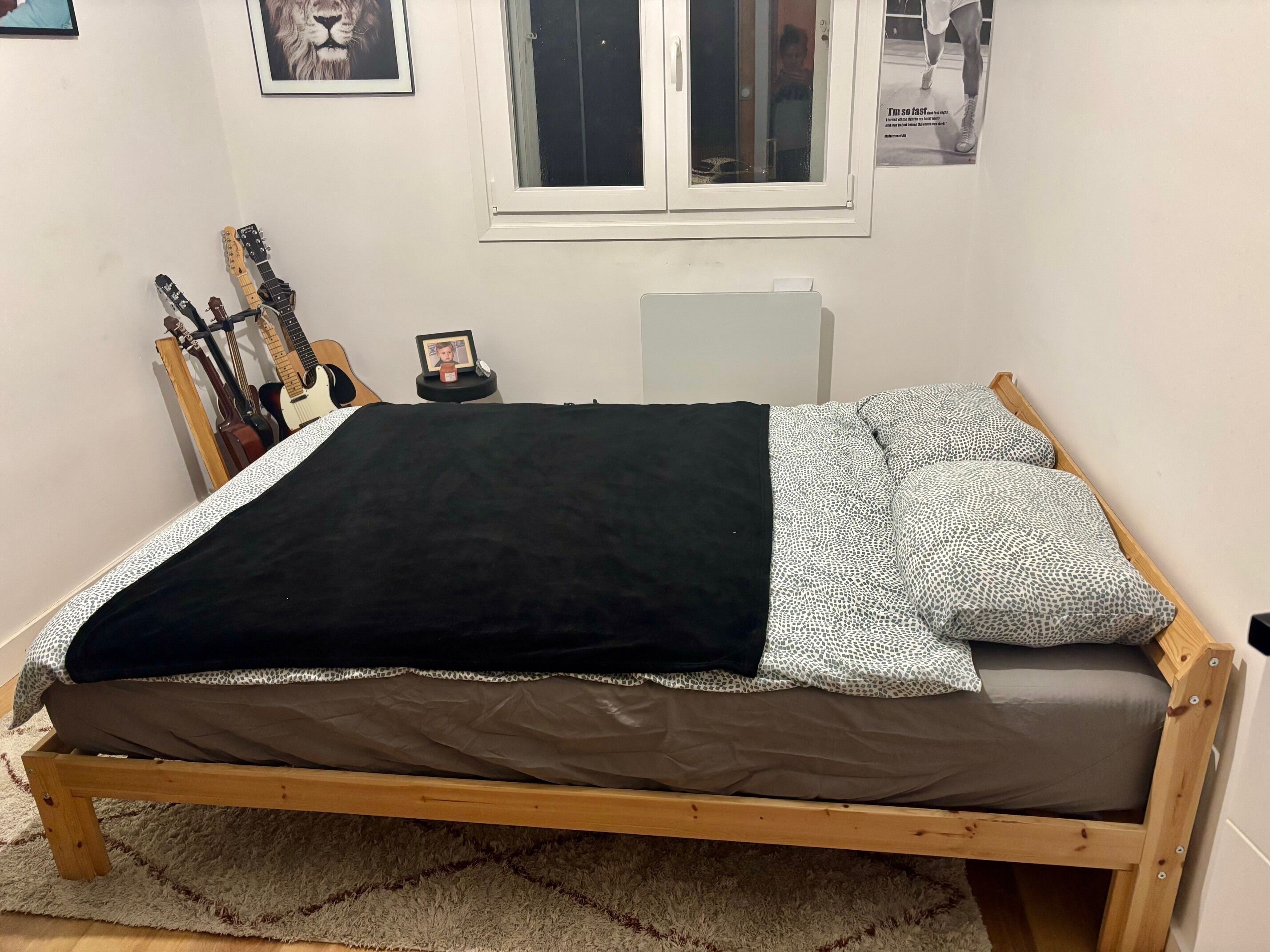 En træseng med et sort og mønstret tæppe, et lille natbord med et foto, og guitarer op ad væggen.