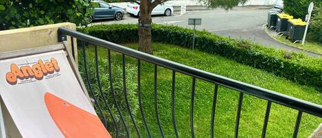 A balcony with a table and a chair, a poster, and a view of a street with cars and greenery.